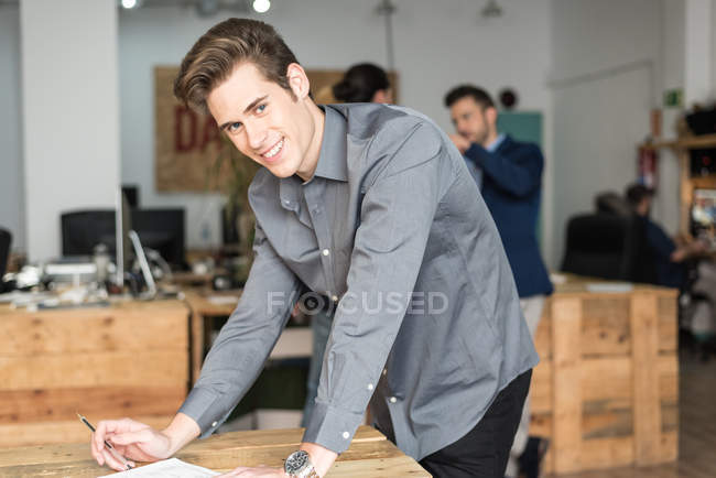 Веселий молодий бізнесмен спирається на стіл з документами і дивиться на камеру — стокове фото