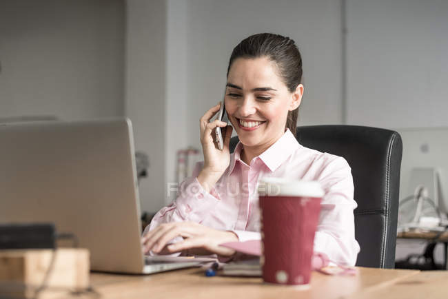 Cheerful brunette businesswoman talking on phone at workplace — Stock Photo