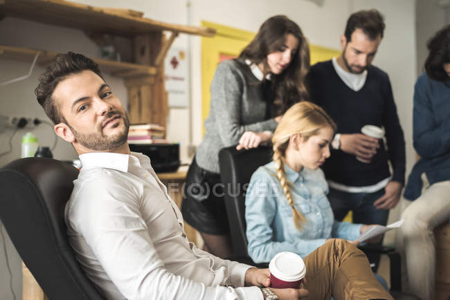 Боковой вид бизнесмена, смотрящего на камеру на фоне людей, работающих в офисе — стоковое фото