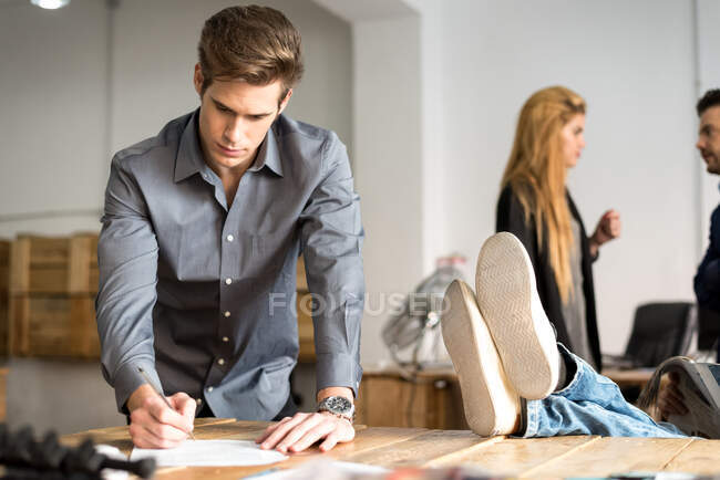 Confident young man signing papers in the office while people doing daily goings. — Stock Photo