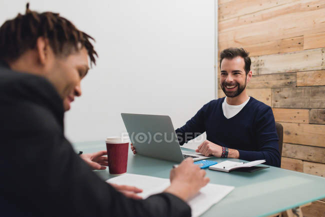 Retrato de hombres de negocios sonrientes sentados a la mesa con laptop y papeles en la oficina - foto de stock