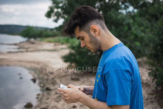 Vista lateral del hombre navegando teléfono inteligente de pie en la costa del lago. Horizontal al aire libre tiro. - foto de stock