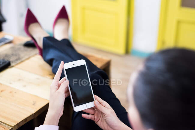 Rückansicht einer Frau, die an ihrem Arbeitsplatz Beine auf den Schreibtisch legt und am Telefon surft. — Stockfoto