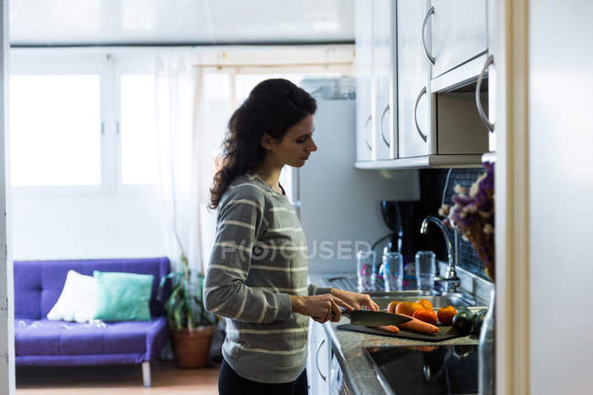Side view of woman with knife cutting carrot on board in kitchen — Stock Photo