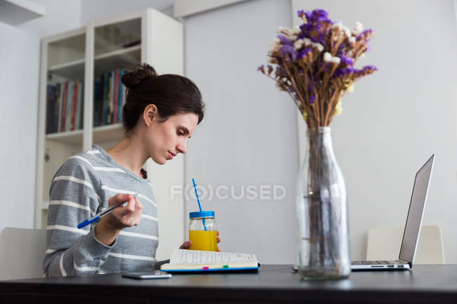 Vista lateral da mulher morena com jarra de suco de laranja sentada à mesa com laptop e olhando para baixo no notebook — Fotografia de Stock