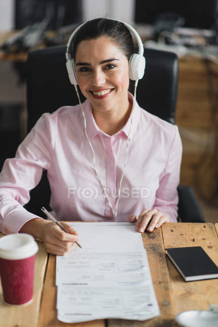 Porträt eines lächelnden Arbeiters mit Kopfhörer, der in die Kamera schaut und am Arbeitsplatz Papiere unterschreibt — Stockfoto