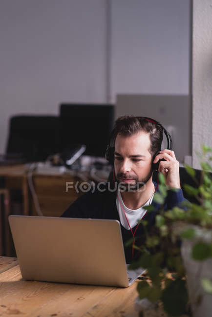 Ritratto di uomo seduto a tavola che aggiusta le cuffie e guarda giù il portatile — Foto stock