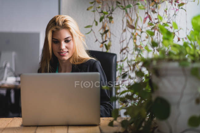 Joven mujer rubia sonriente sentada en la mesa y escribiendo portátil en la oficina . - foto de stock