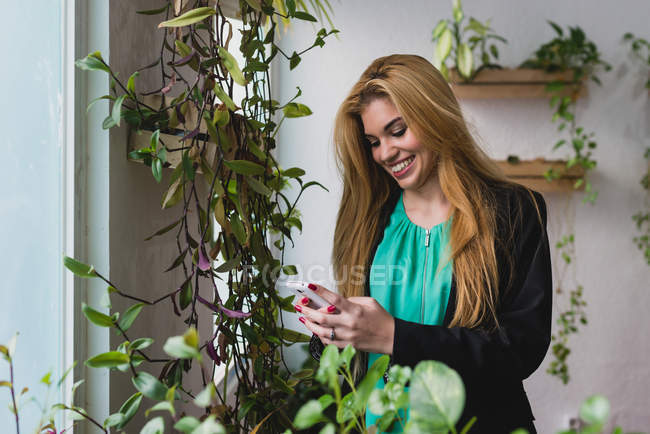 Retrato de chica rubia sonriente de pie cerca de la ventana con plantas en maceta y charlando en el teléfono inteligente - foto de stock