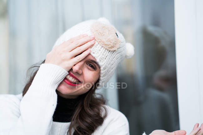 Портрет усміхненої жінки в трикотажному капелюсі, що закриває одне око рукою і дивиться на камеру — стокове фото