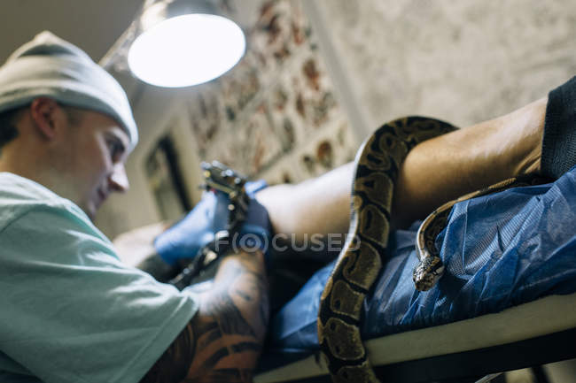 Grande cobra apertando em uma perna enquanto mestre fazendo tatuagem — Fotografia de Stock
