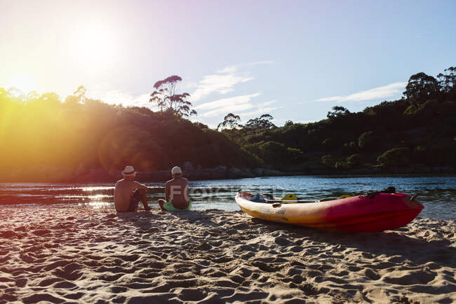Вид ззаду на двох чоловіків, що сидять на піщаному березі з човном і дивляться на сонячне світло . — стокове фото