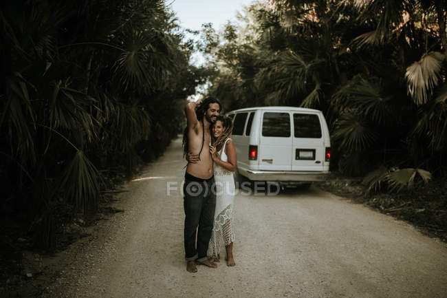 Retrato de pareja con rastas abrazándose en camino de bosque tropical con furgoneta estacionada - foto de stock