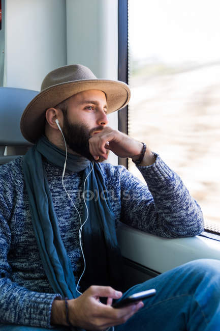 Thoughtful bearded man sitting in train and listening music with earphones — Stock Photo