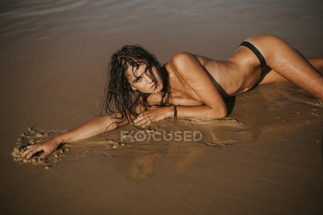 Retrato de mulher de topless com cabelo molhado deitado na praia de areia e olhando para a câmera — Fotografia de Stock