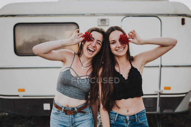 Smiling girls holding red flowers in front of eyes by white trailer car — Stock Photo
