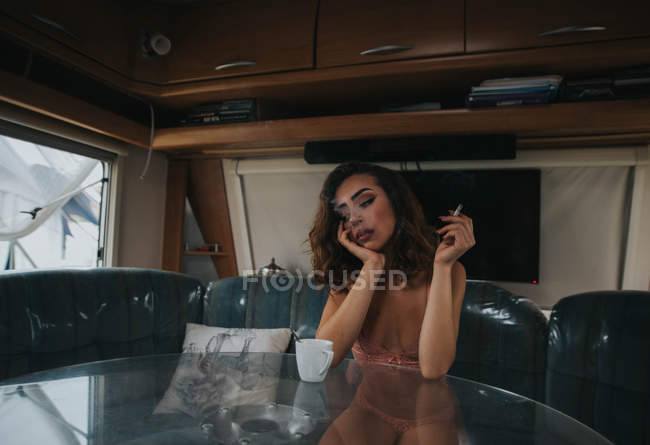 Mujer en ropa interior sentada a la mesa y fumando - foto de stock
