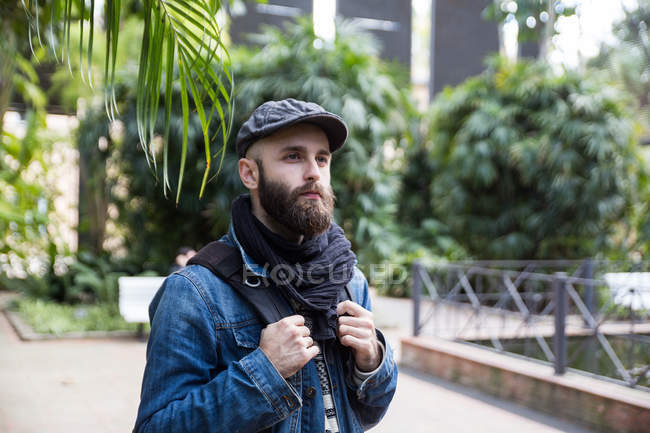 Портрет бородатого чоловіка в стильному вбранні з рюкзаком, який позує на тлі вуличної сцени . — стокове фото