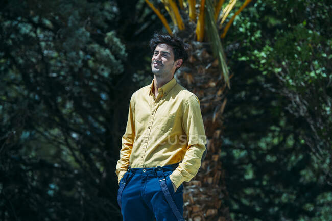 Portrait of confident smiling man in yellow shirt and blue trousers with suspenders smiling away against of trees in sunlight.Holding hands in pockets. — Stock Photo