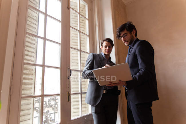 Два молодых сотрудника в костюмах стоят у окна и внимательно смотрят на ноутбук . — стоковое фото