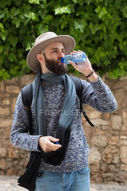 Bearded man in hat with backpack drinking water at street — Stock Photo