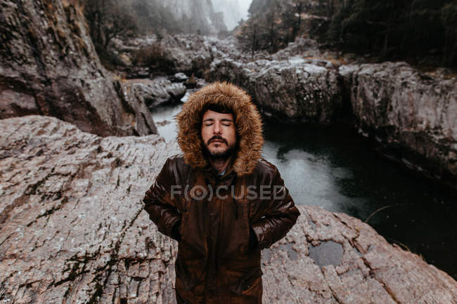 Бородатый мужчина стоит с закрытыми глазами и руками в карманах на фоне горной реки — стоковое фото