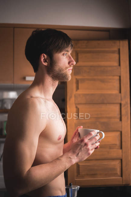 Joven hombre sin camisa sosteniendo la taza y mirando en las ventanas de la cocina - foto de stock