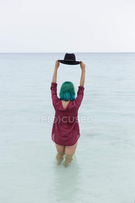Menina sexy irreconhecível em camisa vermelha em pé no mar e segurando chapéu preto acima de sua cabeça de cabelo azul. — Fotografia de Stock