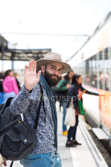 Portrait of bearded man in hat carrying backpack and waving hand at camera over people getting in train on background — Stock Photo