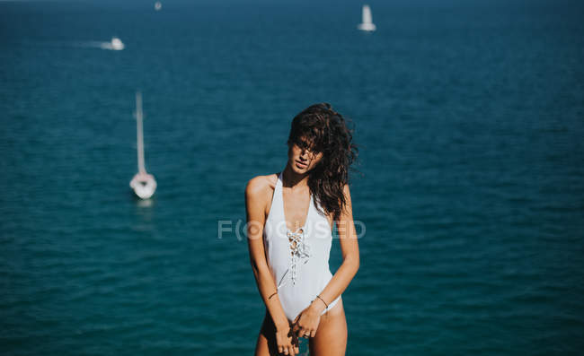 Porträt einer brünetten Frau im weißen Badeanzug, die in die Kamera blickt und mit schwimmenden Yachten gegen den Ozean posiert — Stockfoto