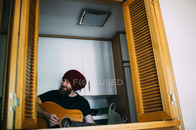 Cheerful bearded man in knitted hat playing acoustic guitar at home — Stock Photo