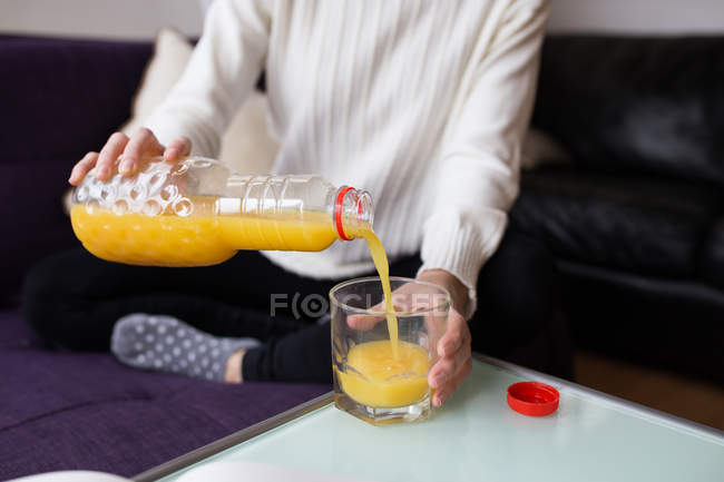 Sección media de la hembra sentada en el entrenador y vertiendo jugo de naranja en el vaso sobre la mesa - foto de stock