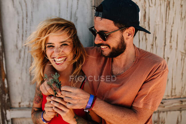 Retrato de la alegre novia rubia riéndose de la cámara mientras se abraza con su novio barbudo en gorra y gafas de sol - foto de stock
