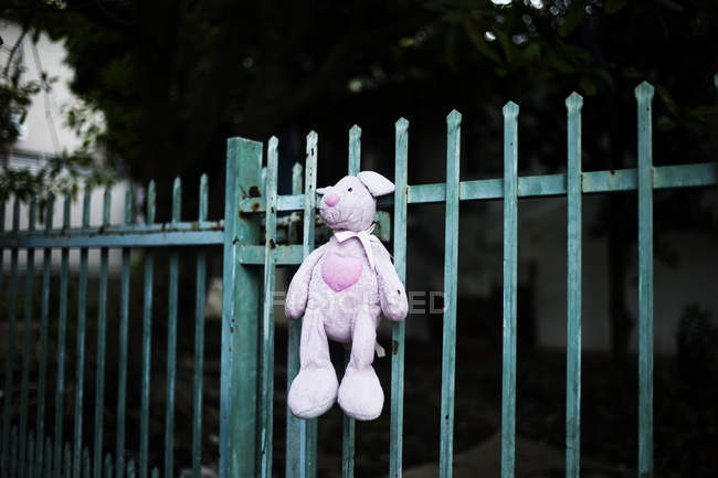 Pink plush rabbit hanging on metal fence with garden yard on background. — Stock Photo