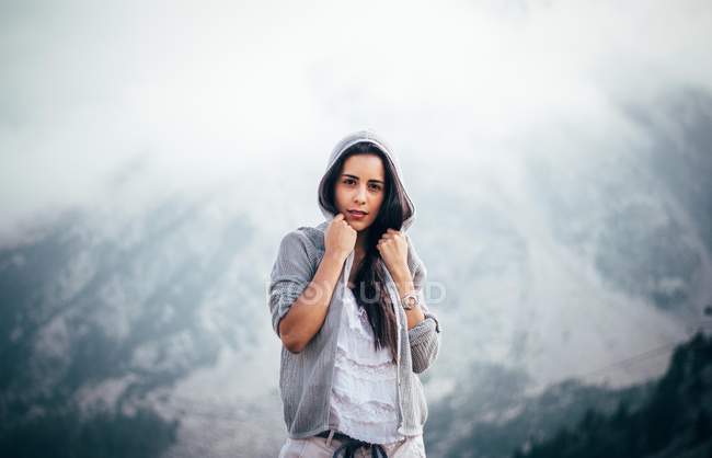 Morena chica en sudadera con capucha posando sobre el paisaje de montaña brumoso - foto de stock