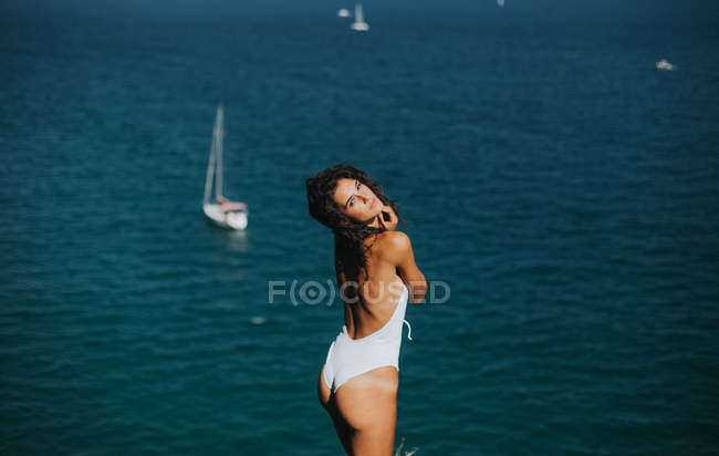 Side view of woman wearing white swimsuit looking to camera and posing against ocean with floating yachts — Stock Photo