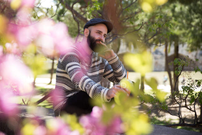 Retrato obscuro de homem barbudo sonhador sentado no parque perto de flores . — Fotografia de Stock