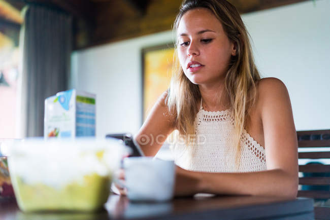 Jeune femme assise et naviguant smartphone tout en prenant le petit déjeuner dans la cuisine . — Photo de stock