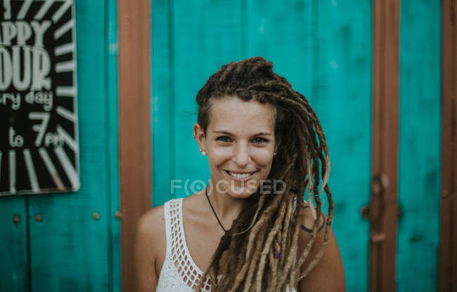 Portrait de femme souriante avec dreadlocks posant sur un mur en bois turquoise et regardant la caméra . — Photo de stock
