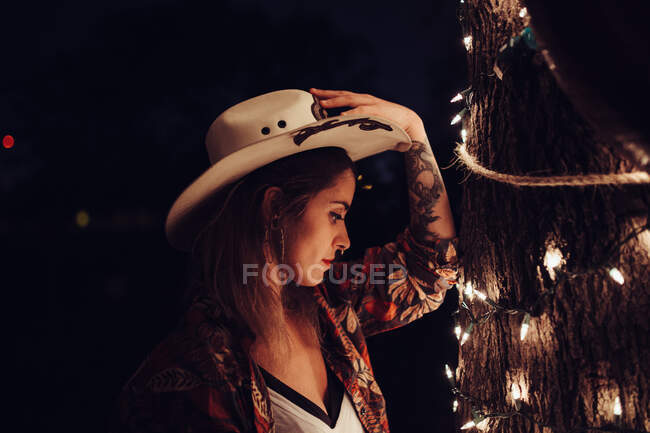 Side view of tattooed stylish woman in cowbow hat standing near tree decorated with burning garland. — Stock Photo