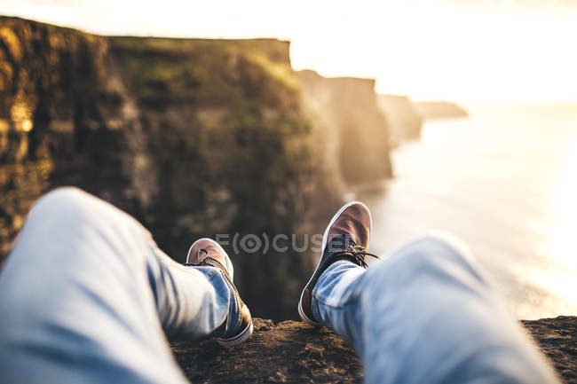 Piernas de hombre con jeans sentados en el acantilado costero - foto de stock