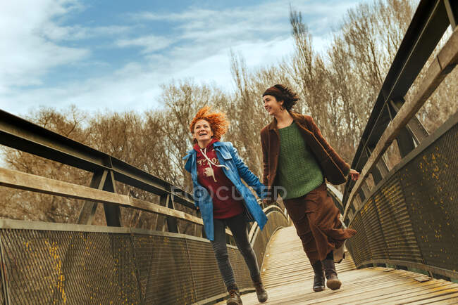 Dos mujeres corriendo por el puente tomadas de la mano. Horizontal al aire libre tiro. - foto de stock