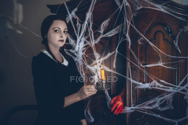 Portrait of plain skin young woman standing next to creepy wardrobe in spiderweb and holding a candle. — Stock Photo