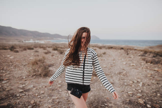 Felice donna bruna con fotocamera digitale sul cinturino a piedi sulla costa del mare — Foto stock