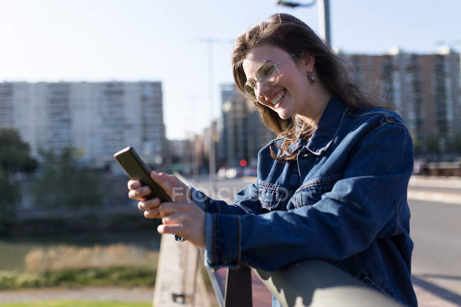 Улыбающаяся женщина, опирающаяся на перила и просматривающая смартфон — стоковое фото