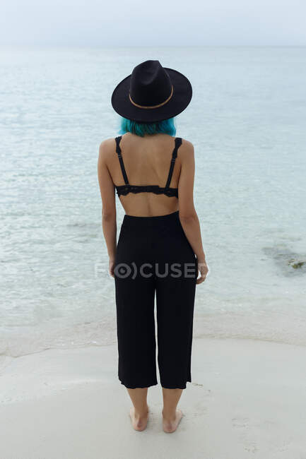 Vista trasera de la irreconocible chica de pelo azul en sujetador negro y pantalones de pie cerca del mar. - foto de stock