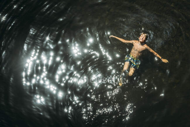 Above view of smiling teen boy relaxing in water with arms outstretched under the sun — Stock Photo