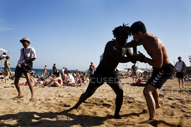 Barcelona, spanien - 10 juli 2011: seitenansicht von zwei jungen männern, die sich am strand prügeln, im hintergrund von menschen, die sich entspannen. — Stockfoto