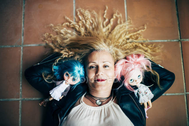 Blonde Frau liegt mit Puppen auf Fliesenboden und blickt in Kamera — Stockfoto