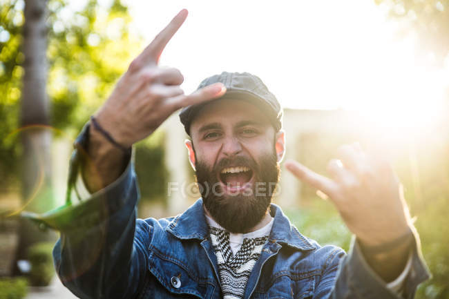 Портрет экспрессивного человека в кепке и джинсовой куртке, кричащего на камеру и жестикулирующего рок-знаком . — стоковое фото